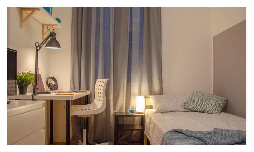 panoramica della stanza con letto singolo, comodino, zona studio con scrivania sedia e lampada, letto singolo